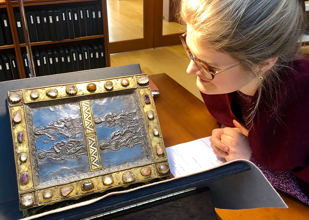 A researcher looking at a large closed manuscript set up in a black and brown book stand on a brown wooden table under a light in the Reading Room of the Morgan Library. The cover of the manuscript is decorated with inlaid stones of different colors in a golden metal border surrounding a blue enamel panel depicting four figures in robes with halos in the center. Visible in the background are some brown wooden shelves with black binders next to glass doors.